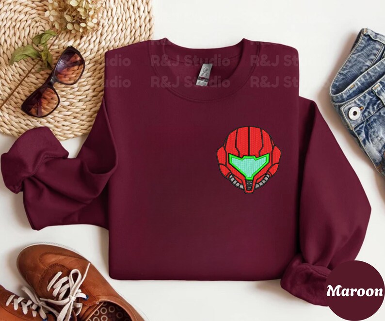 Maroon color unisex crewneck sweatshirt featuring a retro sumas bounty hunter helmet embroidery with red armor panels and neon green visor, minimalist gamer streetwear style
Close up sleeve detail showing embroidered retro sumas helmet patch with red stitching and bright green visor on neutral sand crewneck sweatshirt
