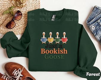 May include: A dark green sweatshirt with the text "Just a Bookish Goose" in orange and green. The design features four cartoon geese reading books. The sweatshirt is paired with brown shoes and blue jeans.