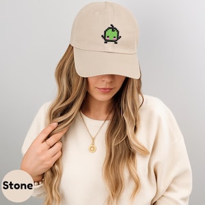 May include: Stone-colored baseball cap featuring a pixelated green and pink cartoon character on the front. The cap has a curved brim and a classic six-panel design. The cap is made of a soft, durable fabric.