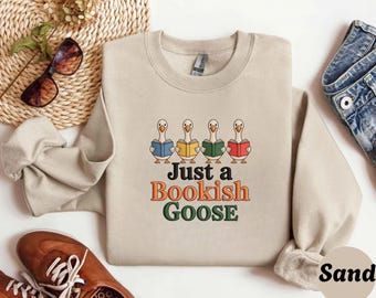 May include: Sand-colored sweatshirt with the text "Just a Bookish Goose" in orange, green, and brown. The design features four cartoon geese, each holding a book with different colored covers. The sweatshirt has a crew neck and long sleeves.