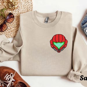 Sand color unisex crewneck sweatshirt featuring a retro sumas bounty hunter helmet embroidery with red armor panels and neon green visor, minimalist gamer streetwear style
Close up sleeve detail showing embroidered retro sumas helmet patch with red stitching and bright green visor on neutral sand crewneck sweatshirt