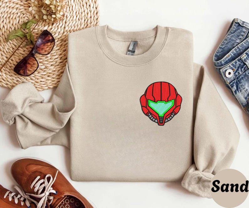 Sand color unisex crewneck sweatshirt featuring a retro sumas bounty hunter helmet embroidery with red armor panels and neon green visor, minimalist gamer streetwear style
Close up sleeve detail showing embroidered retro sumas helmet patch with red stitching and bright green visor on neutral sand crewneck sweatshirt