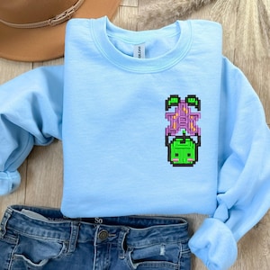 Op de afbeelding: Een lichtblauw sweatshirt met een gepixelde groene en paarse afbeelding. Het sweatshirt is gemaakt van zacht materiaal en heeft lange mouwen. De afbeelding is van een wezen. Het sweatshirt wordt gecombineerd met een blauwe jeans.