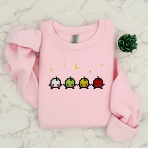 Op de afbeelding: Roze trui met een geborduurd ontwerp van vier gepixelde appels met gezichten, in wit, groen, geel en rood, met een halve maan erboven.