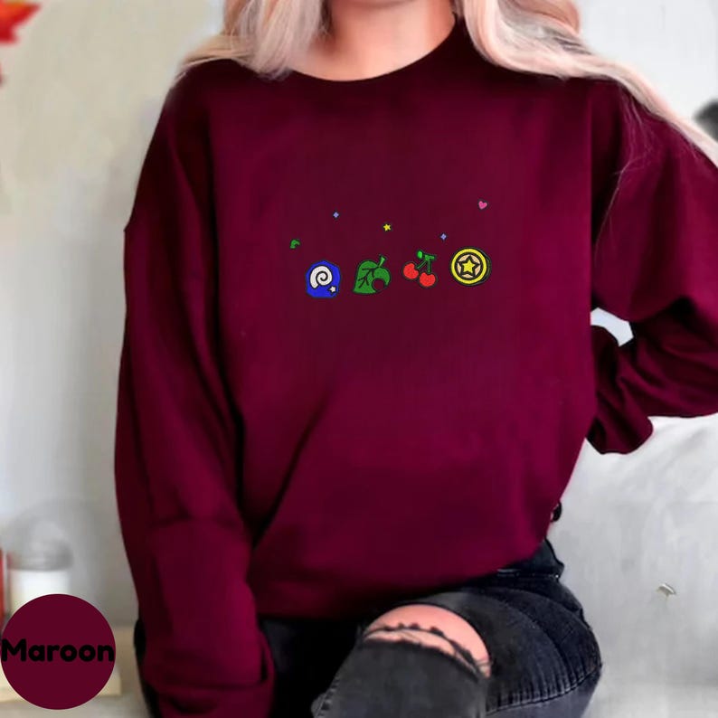 Embroidered Animal Crossing–inspired sweatshirt featuring cute ACNH-style icons stitched on a soft crewneck. Cozy cotton-blend fabric shown in folded and worn views to highlight texture and embroidery detail. Kawaii cottagecore gamer style with pastel color options such as sand, white, blue, and pink. Ideal for cozy gamer outfits, cute embroidered fashion, and cottagecore winter wear. A great gift idea for gamers and fans of soft, comfy embroidered apparel.
