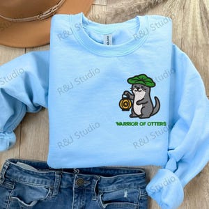 May include: A light blue sweatshirt featuring an embroidered otter wearing a green hat and holding a lantern. The text "WARRIOR OF OTTERS" is below the otter. The sweatshirt is laid on a wooden surface with a pair of blue jeans and a brown hat.