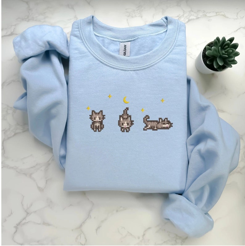 Pu&ograve; includere: Una felpa di colore azzurro chiaro con un design a pixel di tre gatti e una luna crescente. I gatti sono marroni e la luna &egrave; gialla.