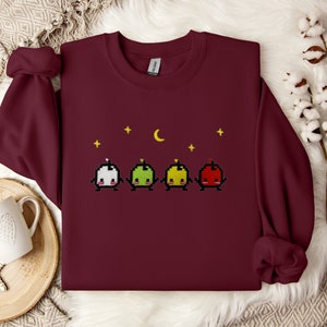 Op de afbeelding: Een bordeauxrood sweatshirt met een pixelontwerp van vier cartoonfiguren met een halve maan en sterren erboven.