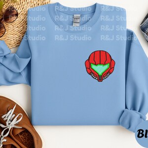 Blue color unisex crewneck sweatshirt featuring a retro sumas bounty hunter helmet embroidery with red armor panels and neon green visor, minimalist gamer streetwear style
Close up sleeve detail showing embroidered retro sumas helmet patch with red stitching and bright green visor on neutral sand crewneck sweatshirt