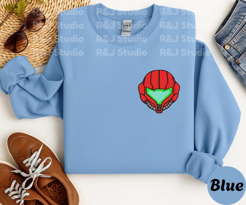 Blue color unisex crewneck sweatshirt featuring a retro sumas bounty hunter helmet embroidery with red armor panels and neon green visor, minimalist gamer streetwear style
Close up sleeve detail showing embroidered retro sumas helmet patch with red stitching and bright green visor on neutral sand crewneck sweatshirt