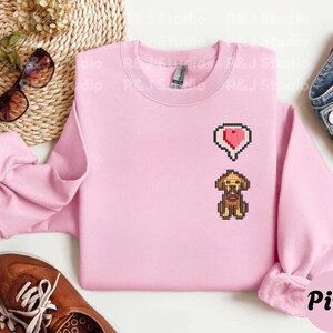 Pink unisex sweatshirt displaying an embroidered Stardew Valley dog in classic pixel art style with a red heart symbol above the character. The soft pastel color combined with the farming game inspired design creates a cozy gamer look, ideal for Stardew Valley fans, casual outfits, relaxed gaming sessions, and cute pixel pet themed apparel.