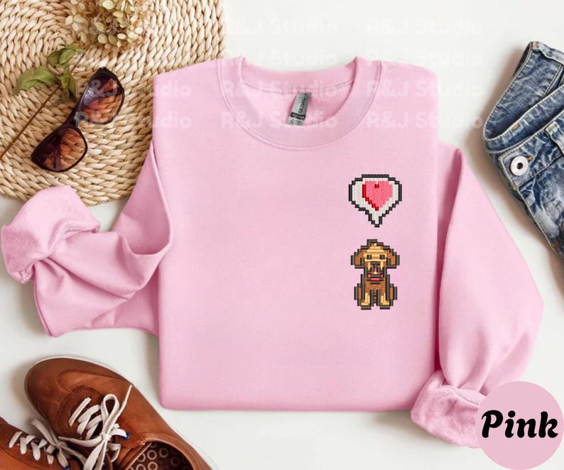 Pink unisex sweatshirt displaying an embroidered Stardew Valley dog in classic pixel art style with a red heart symbol above the character. The soft pastel color combined with the farming game inspired design creates a cozy gamer look, ideal for Stardew Valley fans, casual outfits, relaxed gaming sessions, and cute pixel pet themed apparel.