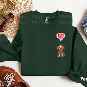 Forest unisex crewneck sweatshirt showcasing a pixel art embroidered Stardew Valley dog with a red heart icon. The contrast stitching enhances the classic farming game aesthetic, making it a cozy gamer apparel choice for Stardew Valley fans, relaxed streetwear looks, indie game inspired outfits, and thoughtful gaming gifts.