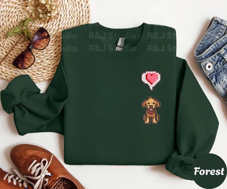 Forest unisex crewneck sweatshirt showcasing a pixel art embroidered Stardew Valley dog with a red heart icon. The contrast stitching enhances the classic farming game aesthetic, making it a cozy gamer apparel choice for Stardew Valley fans, relaxed streetwear looks, indie game inspired outfits, and thoughtful gaming gifts.