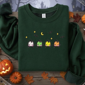 Op de afbeelding: Donkergroene sweatshirt met een gepixeld Halloween-ontwerp. Het ontwerp bevat vier kleurrijke, blokkerige figuren, een maan, sterren en vleermuizen. De sweatshirt is omgeven door Halloween-decoraties, waaronder pompoenen en geesten.