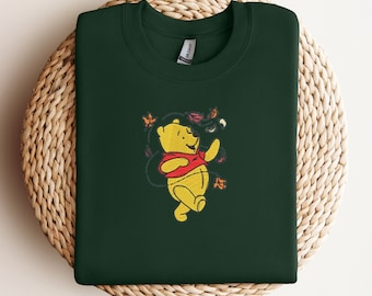 May include: A dark green sweatshirt featuring an embroidered image of a yellow bear wearing a red shirt. The bear is surrounded by leaves. The sweatshirt is folded on a woven, circular surface.