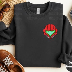 Dark Heather color unisex crewneck sweatshirt featuring a retro sumas bounty hunter helmet embroidery with red armor panels and neon green visor, minimalist gamer streetwear style
Close up sleeve detail showing embroidered retro sumas helmet patch with red stitching and bright green visor on neutral sand crewneck sweatshirt
