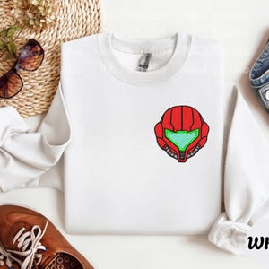 Peut inclure: Sweat-shirt blanc à col rond avec un motif de casque brodé rouge et vert sur la poitrine. Le sweat-shirt a des manches longues et un col côtelé. Le mot "White" est imprimé en bas à droite.