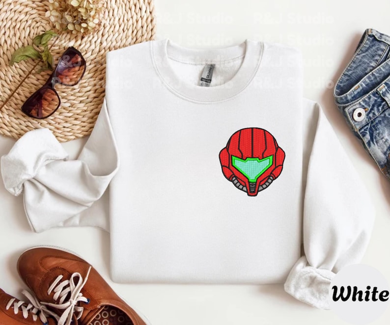 White color unisex crewneck sweatshirt featuring a retro sumas bounty hunter helmet embroidery with red armor panels and neon green visor, minimalist gamer streetwear style
Close up sleeve detail showing embroidered retro sumas helmet patch with red stitching and bright green visor on neutral sand crewneck sweatshirt