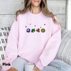 Embroidered Animal Crossing–inspired sweatshirt featuring cute ACNH-style icons stitched on a soft crewneck. Cozy cotton-blend fabric shown in folded and worn views to highlight texture and embroidery detail. Kawaii cottagecore gamer style with pastel color options such as sand, white, blue, and pink. Ideal for cozy gamer outfits, cute embroidered fashion, and cottagecore winter wear. A great gift idea for gamers and fans of soft, comfy embroidered apparel.