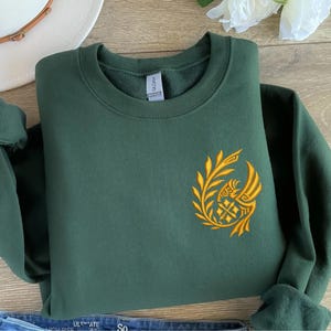 May include: A dark green sweatshirt with a gold embroidered design on the left chest. The design features a stylized bird with a wreath around it.