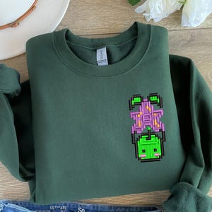 Op de afbeelding: Donkergroene sweatshirt met een gepixeld ontwerp van een groen en paars wezen. De sweatshirt heeft een ronde hals en lange mouwen. Een witte hoed en een blauwe spijkerbroek staan op de achtergrond.