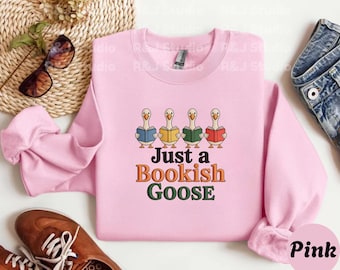 May include: A light pink sweatshirt with the text "Just a Bookish Goose" in orange and green. The design features four cartoon geese reading books. The sweatshirt is a crew neck style.