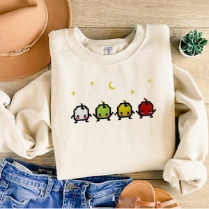 Op de afbeelding: Crèmekleurige sweatshirt met een gepixeld fruitontwerp. Het ontwerp bevat vier verschillende vruchten in verschillende kleuren, met een maan en sterren erboven. De sweatshirt wordt gecombineerd met een blauwe jeans en een beige hoed.