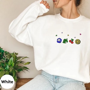 Embroidered Animal Crossing–inspired sweatshirt featuring cute ACNH-style icons stitched on a soft crewneck. Cozy cotton-blend fabric shown in folded and worn views to highlight texture and embroidery detail. Kawaii cottagecore gamer style with pastel color options such as sand, white, blue, and pink. Ideal for cozy gamer outfits, cute embroidered fashion, and cottagecore winter wear. A great gift idea for gamers and fans of soft, comfy embroidered apparel.
