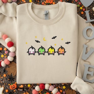 Op de afbeelding: Crèmekleurige sweatshirt met een gepixeld Halloween-ontwerp. Het ontwerp bevat vier kleurrijke pompoenkarakters, vleermuizen, sterren en een halve maan. Een feestelijk en gezellig kledingstuk voor het griezelige seizoen.