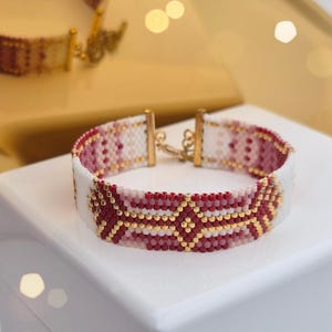 May include: A beaded bracelet featuring a geometric design in red, white, and gold. The bracelet is presented on a white surface, with a second bracelet visible in the background. The clasp is gold-coloured.