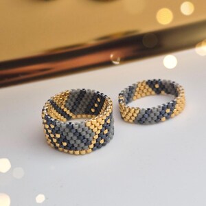 May include: Close-up of beaded rings in shades of gray, gold, and black. The rings are intricately designed with a geometric pattern. The rings are displayed on a white surface with blurred bokeh lights in the background. Jewelry.