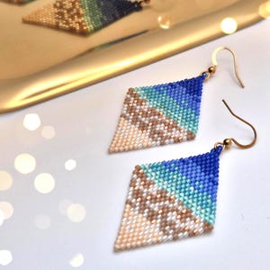 Op de afbeelding: Diamantvormige oorbellen met kralen. De oorbellen hebben een kleurovergang van blauw, turquoise, beige en goud. Ze hebben goudkleurige oorhaken en worden gepresenteerd op een wit oppervlak.