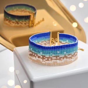 May include: Two beaded bracelets in shades of blue, turquoise, and tan. The bracelets are displayed on a white surface and a gold tray. Each bracelet features gold-coloured metal clasps and a chain.