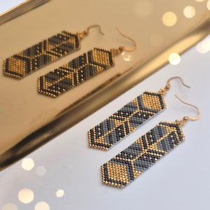 Può includere: Orecchini di perline con un motivo geometrico in oro, grigio e nero. Gli orecchini rettangolari hanno una chiusura a gancio. Gli orecchini sono presentati su un vassoio dorato.