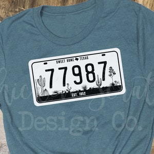 May include: Teal blue t-shirt with a white and black graphic of a Texas license plate that says "Sweet Home Texas" and "77987".