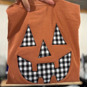 May include: A burnt orange crew neck t-shirt featuring a large jack-o'-lantern face appliqué. The eyes, nose, and jagged mouth are crafted from black and white buffalo plaid fabric, securely stitched onto the front of the garment.