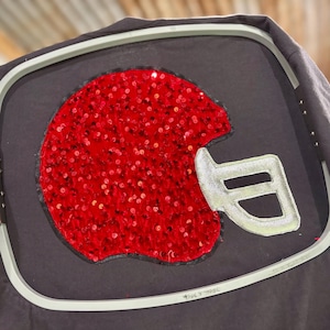 May include: A black t-shirt with a red sequined football helmet design. The helmet has a silver-colored face mask. The shirt is held in a metal embroidery hoop.
