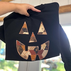 May include: A black long-sleeve sweatshirt featuring a large jack-o'-lantern face applique on the front. The eyes, nose, and mouth are constructed from various quilted fabric patches in shades of orange, brown, cream, and black with stitching.