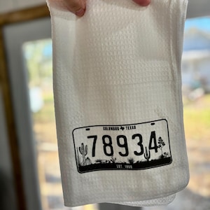 May include: White kitchen towel with a textured pattern. The towel features a black and white graphic of a license plate with the text "COLUMBUS TEXAS" and the number "78934". The bottom of the license plate reads "EST. 1866".