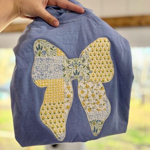 May include: A periwinkle blue sweatshirt featuring a large, central bow appliqué. The bow is constructed from various quilted fabric patches in shades of yellow, white, and blue, displaying floral, gingham, and botanical patterns with visible stitching.