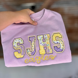 May include: A lilac-colored sweatshirt with the letters "SJHS" in patchwork fabric, featuring floral and gingham patterns. The word "Eagles" is embroidered in yellow script below the letters. The sweatshirt is held up by a hand.