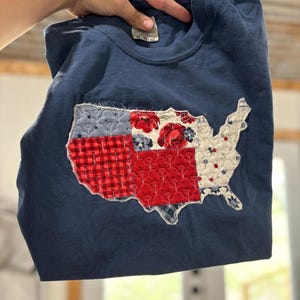 May include: A navy blue long-sleeved shirt featuring a patchwork design of the United States. The patchwork incorporates red, white, and blue fabrics with floral and geometric patterns. The shirt is held up by a hand.