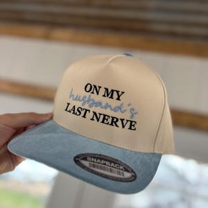 May include: A beige and light blue snapback hat. The hat features the embroidered text "ON MY husband's LAST NERVE" in black and light blue. The brim is light blue, and the hat has a "SNAPBACK" label.