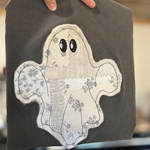 May include: A dark gray t-shirt featuring a large ghost applique. The ghost is constructed from off-white fabric with a subtle floral pattern and a vintage-style text print. It has two large, black embroidered oval eyes and a stitched outline.