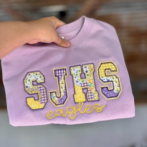 May include: A lilac-colored sweatshirt with the letters "SJHS" in patchwork fabric, featuring floral and gingham patterns. The word "Eagles" is embroidered in yellow script below the letters. The sweatshirt is held up by a hand.