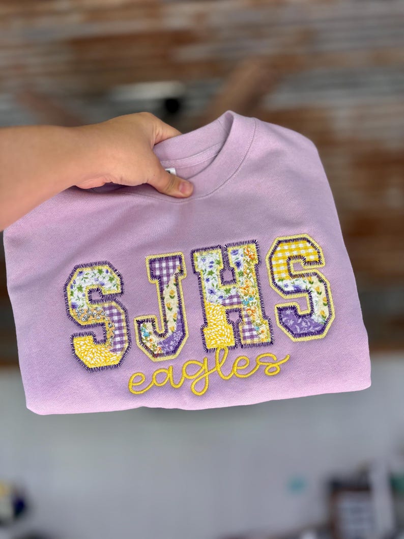 May include: A lilac-colored sweatshirt with the letters "SJHS" in patchwork fabric, featuring floral and gingham patterns. The word "Eagles" is embroidered in yellow script below the letters. The sweatshirt is held up by a hand.