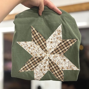May include: Olive green t-shirt with a large starburst design. The starburst is made of alternating plaid and floral fabric patches in shades of brown, cream, and white. The shirt is folded.