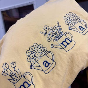 Puede incluir: Una camiseta amarillo claro con regaderas bordadas en azul llenas de flores. Las letras "m" y "a" est&aacute;n bordadas en las regaderas. La camiseta tiene un aspecto suave e informal.