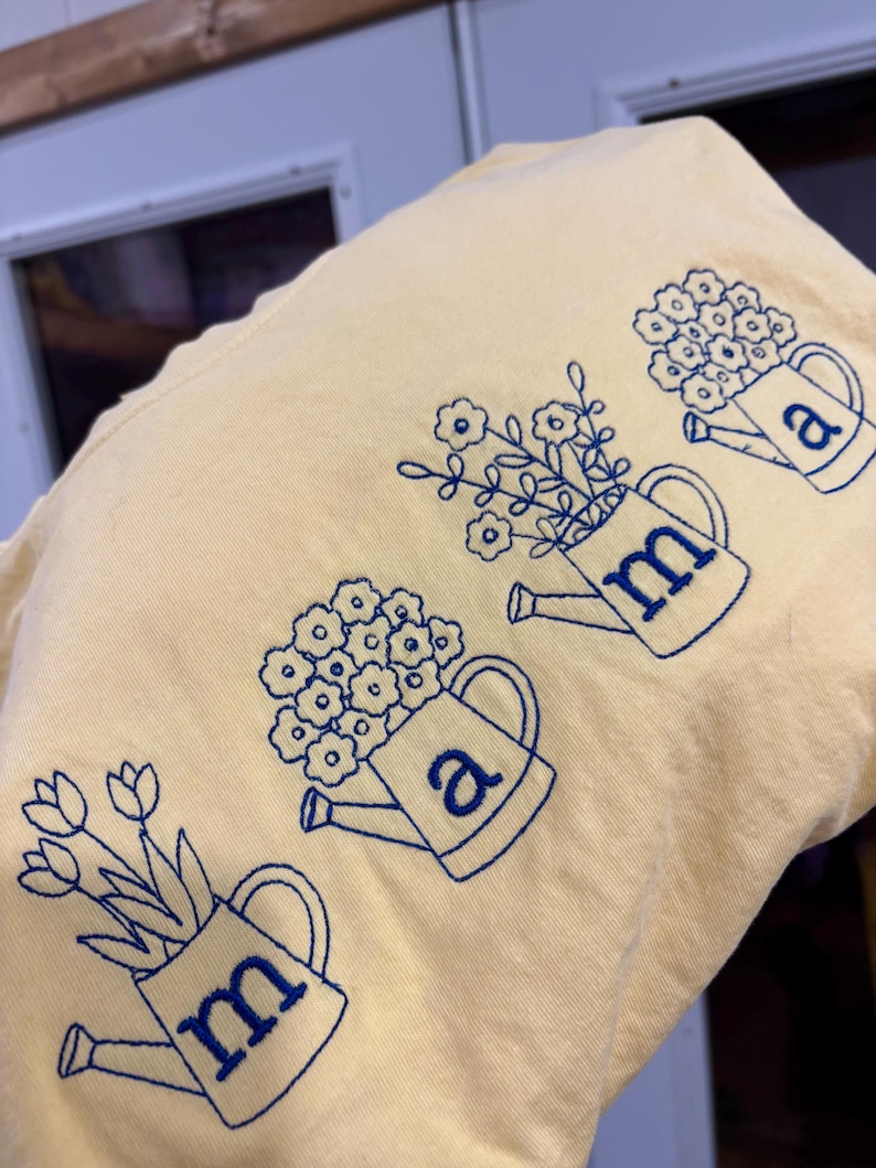 Puede incluir: Una camiseta amarillo claro con regaderas bordadas en azul llenas de flores. Las letras "m" y "a" est&aacute;n bordadas en las regaderas. La camiseta tiene un aspecto suave e informal.